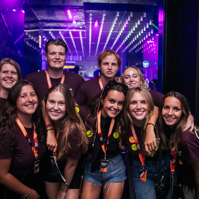 Negen Summer Bash crewleden in België, poserend in een rustige club, allemaal met Summer Bash-shirts en lanyards, klaar om jongerenreizen te begeleiden.